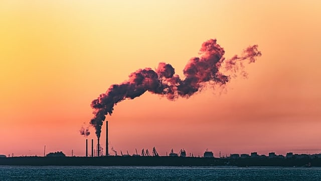 The skyline of a harbor in the evening light, the sky is red-orange. A single chimney emits smoke, the clouds of smoke drift across the sky.