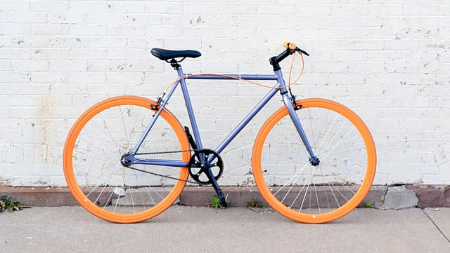 Blaues Fahrrad mit orangenen Reifen vor einer weißen Wand.