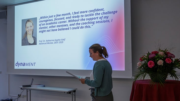 Eine Frau steht vor einer Videoleinwand. Dort sind eine andere Frau und ein Zitat abgebildet.
