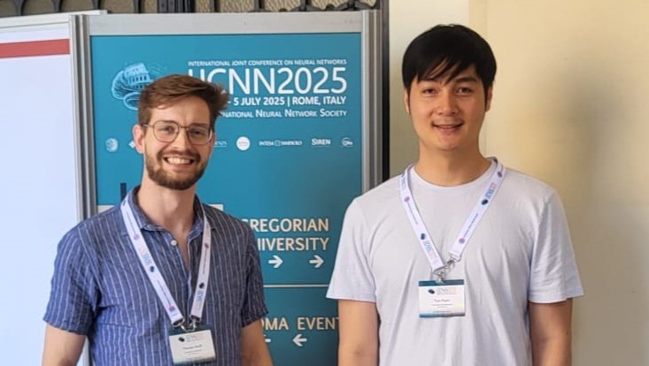 two doctoral candidates standing in front of the ijcnn conference sign