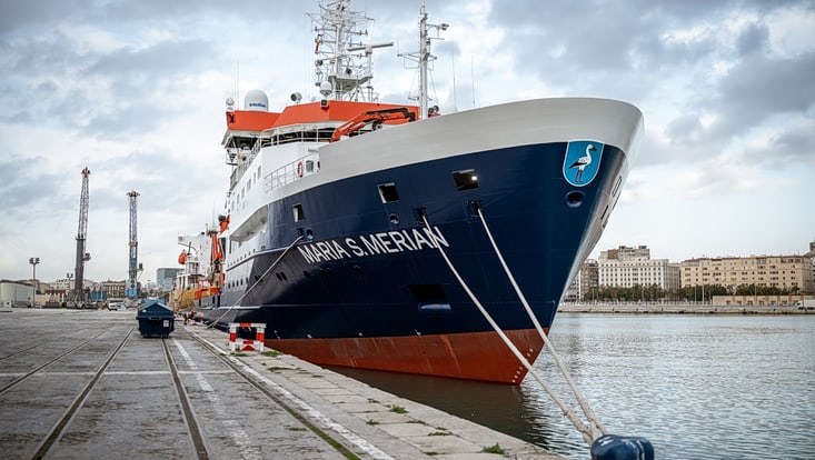 The research vessel Maria S. Merian is moored on the quay with its bow facing the viewer. Its name can be clearly read in white lettering on the dark blue bow.