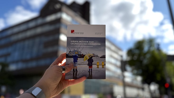 Cover des Booklets "Städte müssen sich grundlegend verändern - Zehn Klimaforscher:innen berichten", im Hintergrund verschwommen das Haus der Erde.