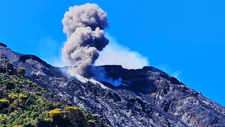Das Bild zeigt eine typische Eruption des Vulkanes. Eine große graue Wolke steigt aus dem Vulkan empor. Der Himmel ist blau, nur eine kleine weiße Wolke ist hinter dem Vulkan zu sehen.