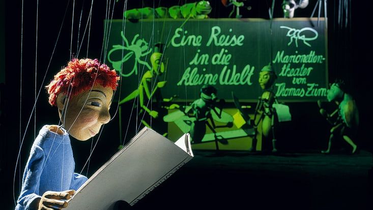 Marionetthe im Vordergrund vor dunkelgrünem Hintergrund. Die Marionette liest in einem großen weißen Buch.