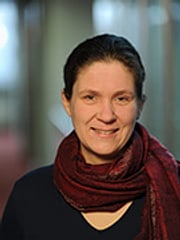 Das Bild ist ein Portrait von Johanna Baehr, einer weißen, weiblich gelesenen Person mittleren Alters. Johanna hat dunkle, zurückgebundene Haare und trägt einen dunkelroten Schal und ein schwarzes Oberteil. Johanna lächelt.
