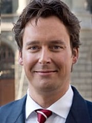 Profilbild von Timo Busch: Eine männlich gelesene Person mit kurzen braunen Haaren. Timo trägt einen dunkelblauen Anzug, ein weißes Hemd und eine rote Krawatte. Timo lächelt.
