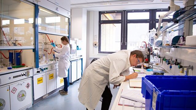 Studentin am Abzug und Student mit Schutzbrille schreibt etwas ins Heft im Labor