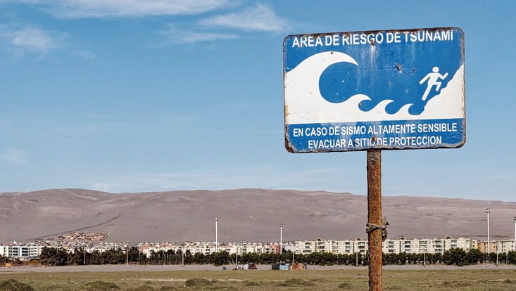 A rusti tsunami evacuation sign at the beach in Arica, Chile. In the background hotels and grey mountains.