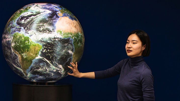 A person is standing in front of a large globe and pointing at it.