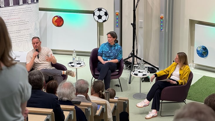 Zu sehen ist ein Hörsaal wo vorne drei Personen sitzen und Mikrofone in den Händen halten.