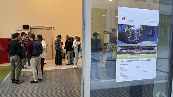 Zu sehen ist eine offene Tür wo an der rechten Seite ein Plakat hängt und im Hintergrunf verschwommen Menschen stehen.