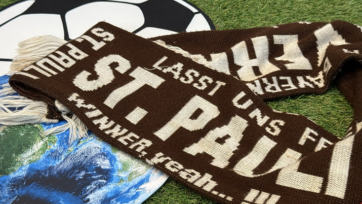 Zu sehen sind eine Erdkugel und ein Fußball auf dem ein St. Pauli Schal liegt.