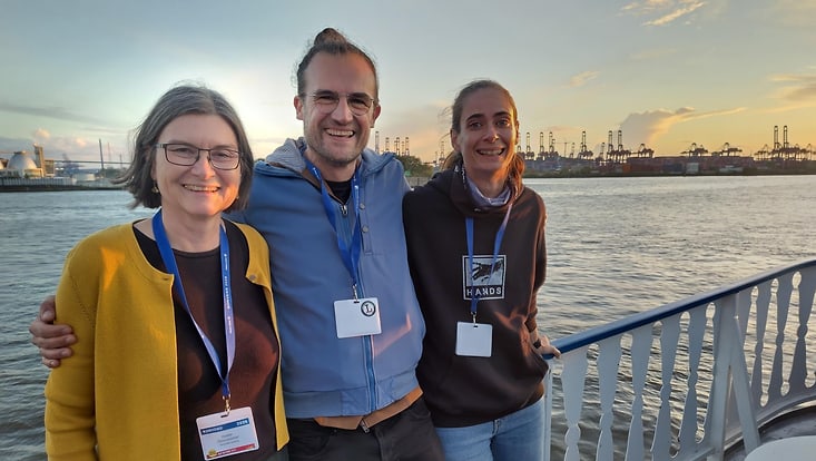Organisatoren Prof. Heike Zinsmeister, Prof. Chris Biemann and Prof. Anne Lauscher lächeln in die Kamera, an Bord eines Ausflugschiffes mit dem Hamburger Hafen im Hintergrund
