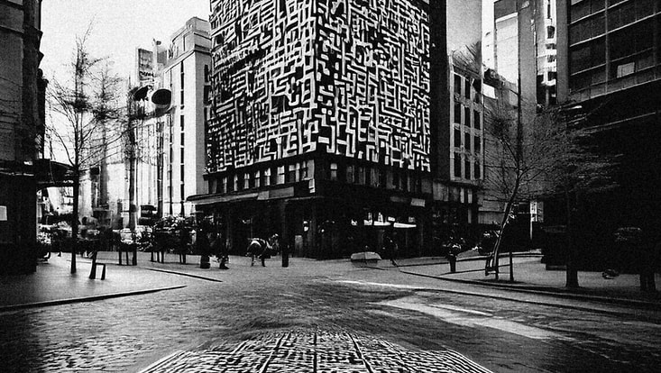The black and white image shows a city scene with a large sky scraper in the middle. Embedded on the large tower building is a QR code pattern in white. In the paved road leading to the block is also a QR code like pattern.