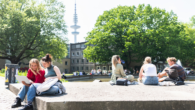 V3A1931_belebter Campus640 Foto vom Ententeich bei sonnigem Wetter, Studierende sitzen in Gruppen draußen und unterhalten sich, essen, schauen auf einen Laptop