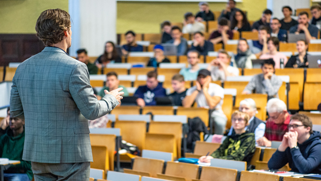 Professor hält eine Vorlesung im Hörsaal, man sieht ihn von hinten. Im Hörsaal sitzen viele Studierende.