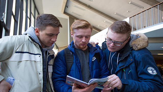 Drei Schüler stehen nebeneinander und schauen in ein Heft, das der Schüler in der Mitte in der Hand hält.