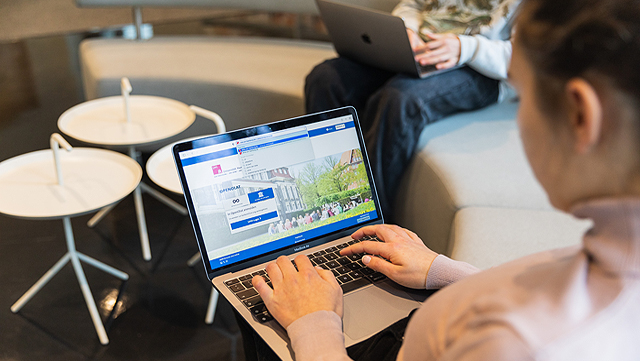 Zwei Personen sitzen mit ihren Laptops auf einem Sofa. Man sieht der einen Person über die Schulter und erkennt den Bildschirm. Auf dem Bildschirm sieht man eine geöffnete Webseite.