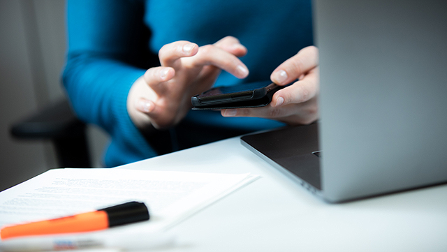 Person in blauem Pullover sitzt vor einem Laptop und hält ein Handy in der Handy. Sie gibt etwas am Handy ein.