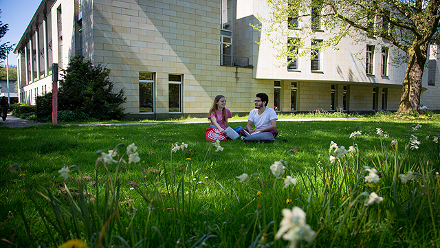 Zwei Studierende sitzen auf einer kleinen Wiese vor einem Unigebäude.