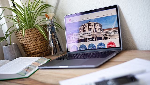 Ein Laptop steht auf einem Schreibtisch. Neben dem Laptop steht eine Pflanze und davor liegt ein aufgeschlagenes Buch.