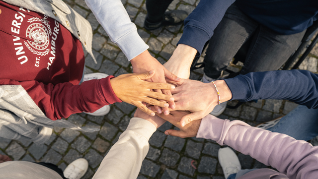 Foto von oben aufgenommen, mehrere Personen legen ihre Hände in der Mitte aufeinander.