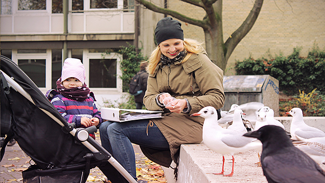Mutter sitzt auf einer Betonbank. Auf ihrem Schoß liegt ein Ordner. Rechts von ihr sitzen ein paar Möwen. Links von ihr steht ein kleines Kind und ein Kinderwagen-