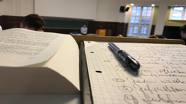 Foto von einem Notizzettel und einem offenen Buch, aufgenommen in einem Hörsaal, aus Sicht eines/einer Studierenden.