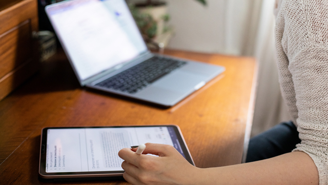 Eine Person sitzt am Schreibtisch, vor ihr steht ein Laptop und sie schreibt etwas auf ihrem Ipad.