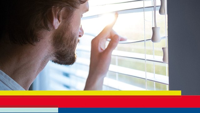 Mann schaut durch Jalousie aus Fenster