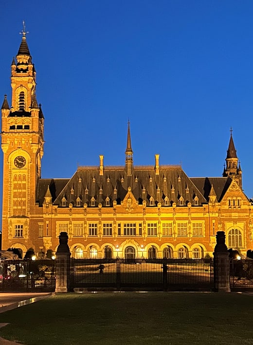 Das Bild zeigt den Friedenspalast in Den Haag bei Nacht, dramatisch beleuchtet vor einem tiefblauen Himmel. Links dominiert der hohe Uhrenturm mit seiner spitzen, dunklen Kuppel das Foto - die große, beleuchtete Uhr ist deutlich sichtbar.