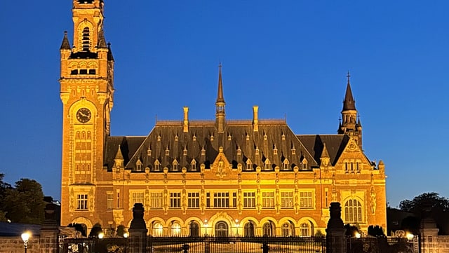 Das Bild zeigt den Friedenspalast in Den Haag bei Nacht, dramatisch beleuchtet vor einem tiefblauen Himmel. Links dominiert der hohe Uhrenturm mit seiner spitzen, dunklen Kuppel das Foto - die große, beleuchtete Uhr ist deutlich sichtbar.