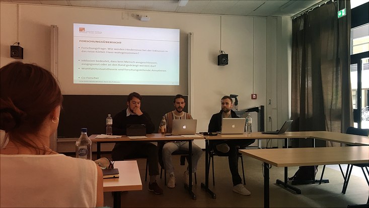 3 Studierende stellen sitzend Ergebnisse zum Thema "Inklusion in das neue Körber-Haus" im Seminarraum vor