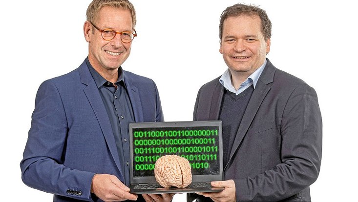 Prof. Dr. Christian Gerloff (links) und Prof. Dr. Martin Spindler halten einen Laptop, auf dessen Tastatur das Modell eines Gehirns tront. Auf dem Bildschirm ist eine Folge von grünen Nullen uns Einsen zu sehen.