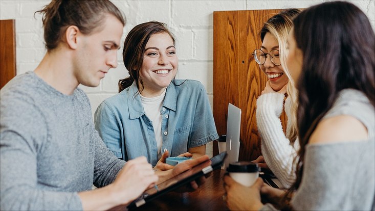 Vier Studenten sitzen lächelnd um einen Tisch, trinken Kaffee und arbeiten an Laptops und Tablets