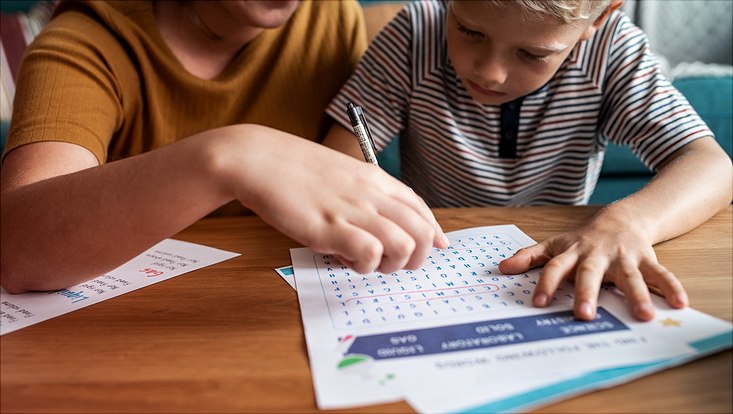 Person helping child to fill in homeschooling work sheet on kitchen table