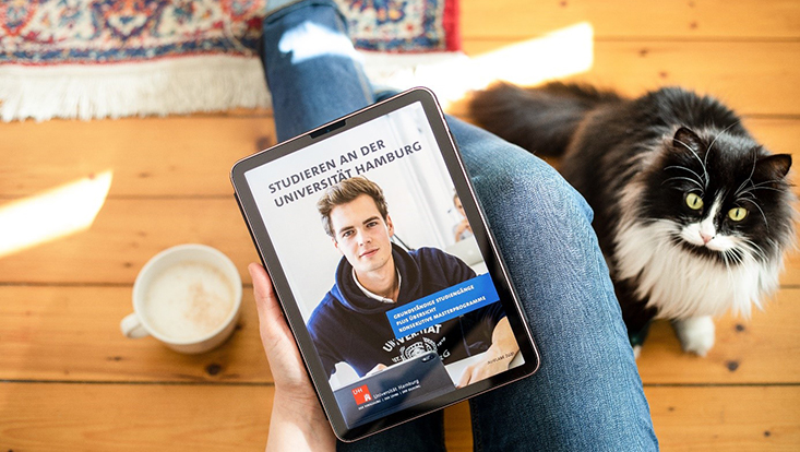 Ein Student sitz auf dem Holzboden neben einer Katze und hält ein Tablet in der Hand, auf dem die Seite "Studieren an der Universität Hamburg" geöffnet ist
