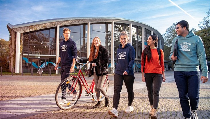 Studis auf dem Campus. Die Frau mit rotem Haar schiebt ein rotes Damenrad.