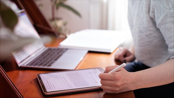 Weibliche Person sitzt an einem Holztisch mit Laptop und Tablet