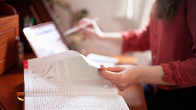 weibliche Person mit rotem Hemd vor einem Holztisch mit Buch und Laptop