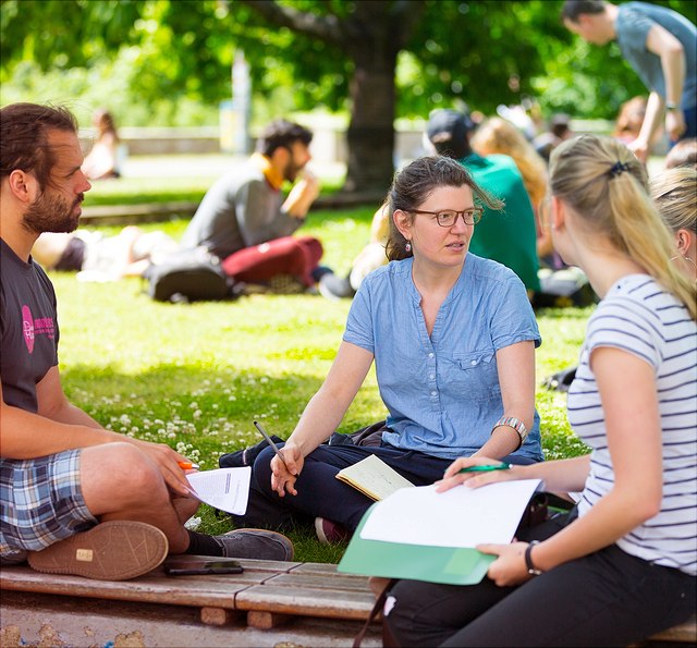 Ein männlicher und drei weibliche Studierende sitzen auf der Wiese und reden