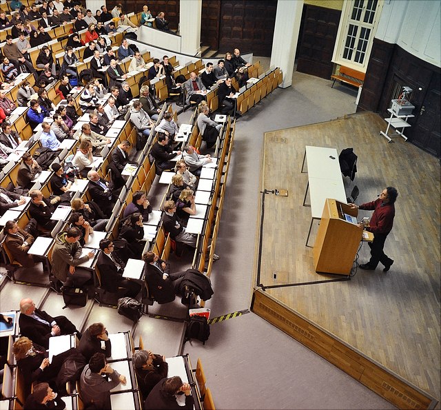 Hörsaal mit vielen Studierenden und männlichen Lehrenden