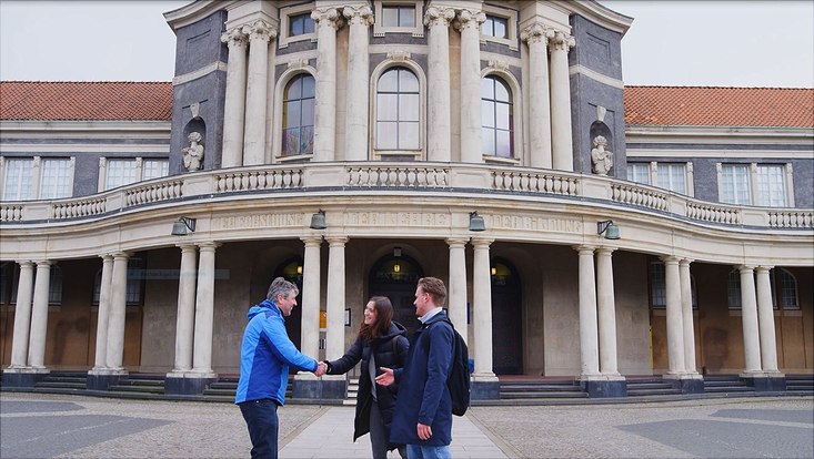 Dieses Bild zeigt das Hauptgebäude der Uni Hamburg und drei Personen die sich begrüßen