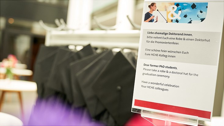 Dieses Bild zeigt einen Wegweiser von der Promoviertenfeier. Im Hintergrund sieht man schwarze Roben und Hüte.