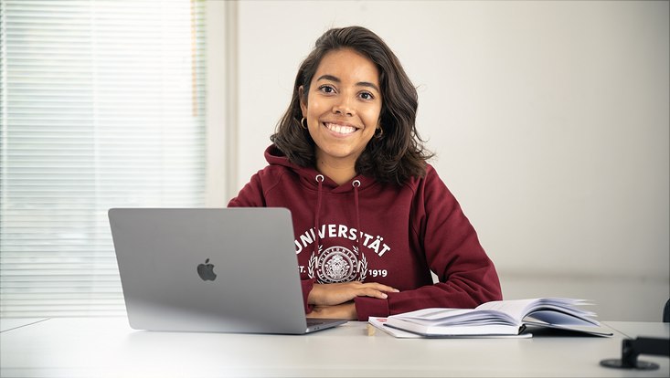 Studentin, die am Tisch sitzt. Vor ihr ein Laptop und einige Bücher.