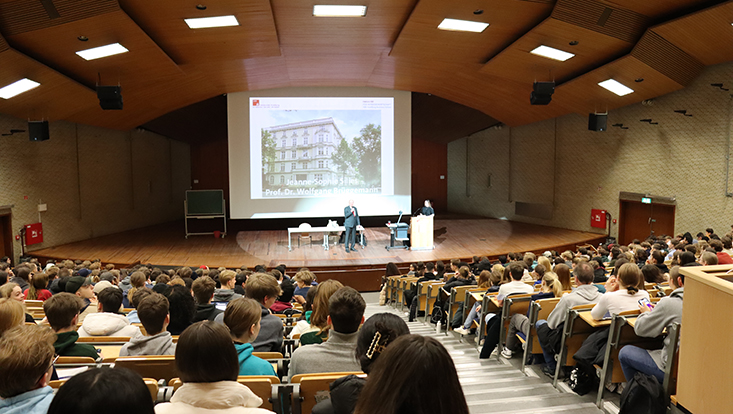 Audimax, Foto aus den hinteren Reihen mit Blick zur Bühne aufgenommen, auf der Prof. Brüggemann seinen Vortrag hält.