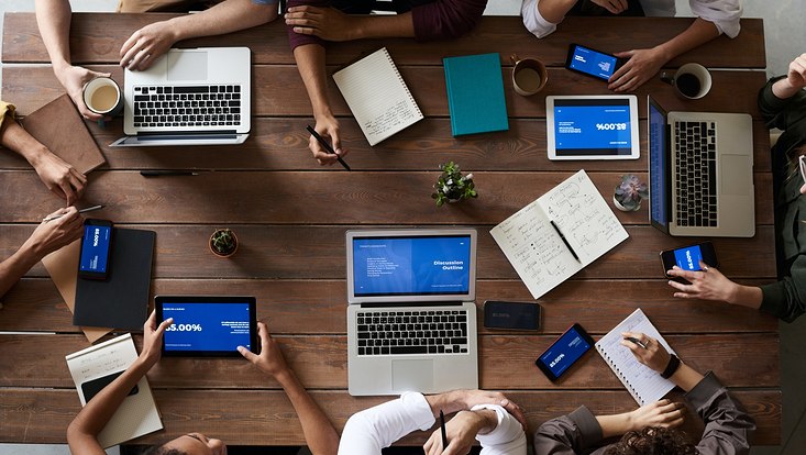 Einige Laptops stehen aufgeklappt auf einem Tisch, mehrer Leute sitzen um den Tisch herum und arbeiten.