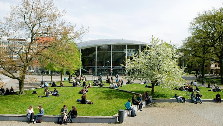 Blick auf das Audimax der UHH, im Vordergrund Studierende auf der Wiese, Bäume mit Frühlingsblüten