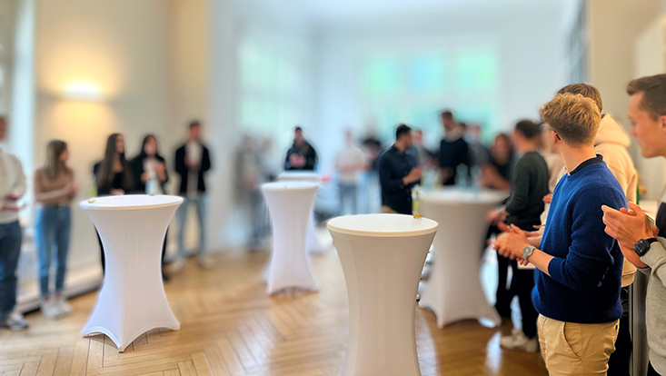 Im Konferenzraum mit Holzfußboden und großen Fenstern stehen Studierende an Stehtischen mit weißen Hussen.
