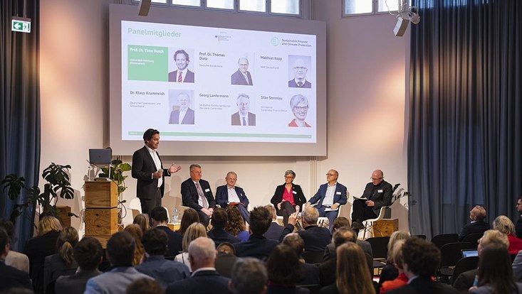 Prof. Busch steht auf einer Bühne und moderiert eine Paneldiskussion vor Publikum